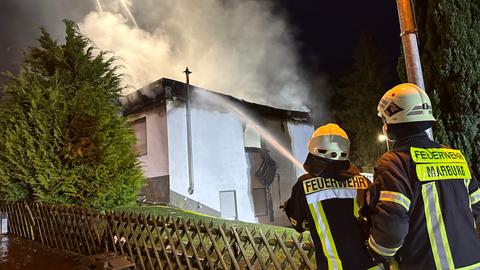 Feuerwehrleute löschen mit einem Schlauch. Aus einem Haus steigt Rauch. Es ist noch dunkel.