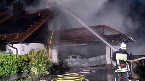 Ein Feuerwerhmann richtet seinen Wasserschlauch auf ein zweistöckiges Einfamilienwohnhaus. Daraus und aus der angrenzenden Garage qualmt nach einem Brand dichter Rauch. In der Garage steht ein weißes Auto. Das Foto wurde nachts aufgenommen.