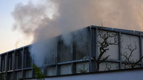 Das Bild zeigt eine rechteckige Lagerhalle mit Flachdach. Aus den mit Metallstreben verkleideten Fenstern dringt grauer Rauch.