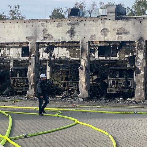 A man walks along in front of the burnt down equipment shed, with hoses lying on the ground.