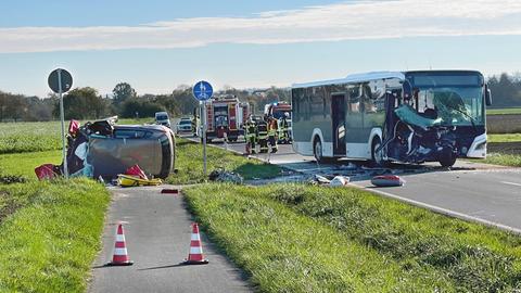 Ein zerbeultes Auto liegt auf einem Radweg, ein Bus mit stark zertrümmerter Front steht auf der Straße, daneben Polizei und Rettungskräfte