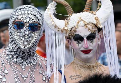 Teilnehmende am CSD: Eine Person mit Strass-Stein-Gesichtsmaske und blauer Brille, eine Person mit Horn-Kopfputz und Goldschmuck