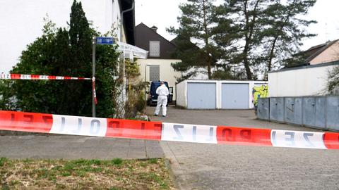 Rot-weißes Flatterband mit dem Aufdruck POLIZEI, dahinter ein Wohnhaus und leicht unscharf eine Person der Spurensicherung vor dem Hauseingang.