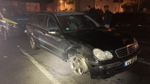 Bei einem stehenden schwarzen Mercedes ist nur noch die Felge am Vorderrad zu sehen, kein Reifen mehr. Im Hintergrund ein Polizeiwagen.