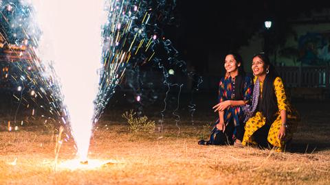 Zwei Frauen knien nebeneinander auf dem Boden und schauen ein fontäneartiges Bodenfeuerwerk vor ihnen an.