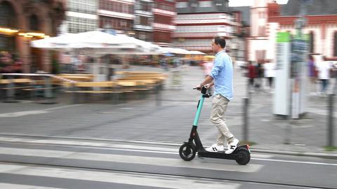 Ein Mann auf einem E-Scooter währen der Fahrt auf einer Straße. Im Hintergrund unscharf ein städtischer Platz.