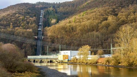 Ein Kraftwerksgebäude am Fluss neben dem sich Hochdruckrohre den Berg hinaufschlängeln