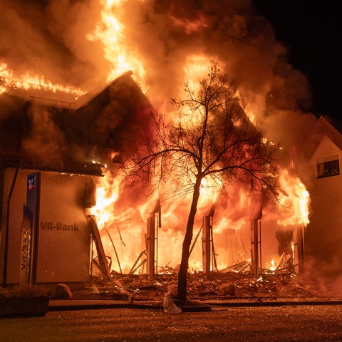 Flammen schlagen aus einem Bankgebäude, davor liegen Trümmer und Splitter