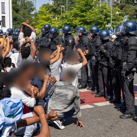 Menschen mit verpixelten Gesichtern, Fäusten in der Luft, manche am Boden, vor einer Polizeikette.