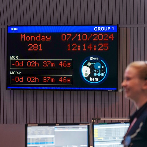 Blick in das Steuerungszentrum der ESA zum Start der "Hera"-Mission im All, digitale Anzeigetafel mit Uhrzeit und Datum, lächelnde Mitarbeiter