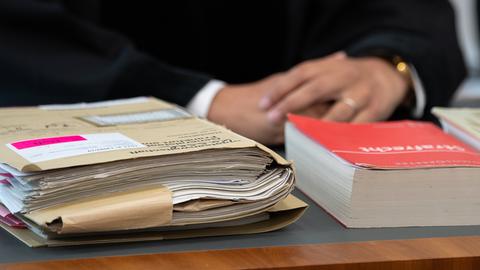 Auf einem dunklen Holztisch liegt eine dicke Akte, daneben ein in rot eingebundesenes Strafgesetzbuch. Im Hintergrubnd sieht man die gefalteten Händes eines Richters.