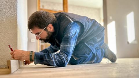 Ein Mann in Arbeitskleidung hält eine Fußleiste aus Holz an eine Wnad. Mit dem Bleistift zeichnet er die richtige Länge ein.