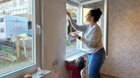 Eine Frau steht auf einer Leiter und spachtelt die Laibung an einem geöffneten Fenster. Unten steht ein kleines Mädchen und schaut zu.