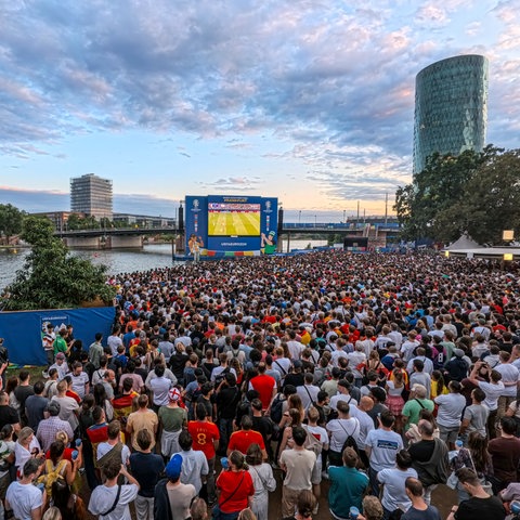 Fans stehen vor dem Big Screen auf der Fanzone, im Hintergrund ist der Main zu sehen. 