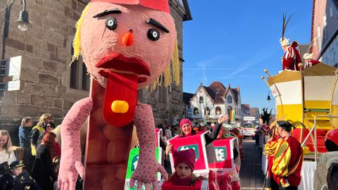 Rosenmontagszug in Volkmarsen: Ein Wagen mit einer überlebensgrpßen Puppe aus Pappmaschee mit einer langen roten Zunge und einem gelben Bonbon darauf. Dahinter gehen Menschen, die sich als TicTac-Bonbonschachteln verkleidet haben.