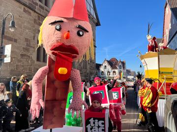 Rosenmontagszug in Volkmarsen: Ein Wagen mit einer überlebensgrpßen Puppe aus Pappmaschee mit einer langen roten Zunge und einem gelben Bonbon darauf. Dahinter gehen Menschen, die sich als TicTac-Bonbonschachteln verkleidet haben.