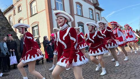 Beim Rosenmontagszug in Fritzlar war eine Gruppe Gardemädchen in rot-weißen Kostümen und rot-weißen dreieckigen Hüten vor einem historischen Gebäude unterwegs. Die Sonne scheint.