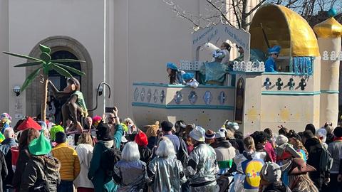 Beim Rosenmontagszug in Seligenstadt war der Wagen des Prinzenpaars zu sehen: Auf einem Wagen waren mehrere halboffene Kuppeln hintereinander montiert. Auf dem Balkon der vordersten stehen Menschen in hellblauen Kostümen. Davor ein Wagen mit einer Affenfigur und einer Palme. Viele Menschen am Straßenrand schauen zu.