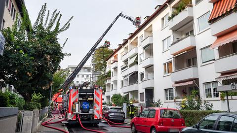 Ein Feuerwehrauto steht in einer städtischen Straße vor einem Mehrfamilienhaus und hat eine Leiter zum Dach ausgefahren, aus welchem Rauch aufsteigt.