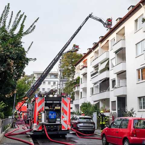 Ein Feuerwehrauto steht in einer städtischen Straße vor einem Mehrfamilienhaus und hat eine Leiter zum Dach ausgefahren, aus welchem Rauch aufsteigt.