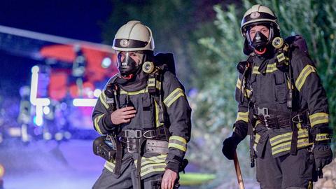 Im Bildvordergrund zwei Feuerwehrleute in voller Montur mit Atemschutzmasken. Im Hintergrund - unscharf - eine Landstraße in der Dämmerung und Feuerwehrauto im Einsatz.