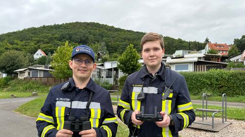 Zwei Feuerwehrmänner stehen mit Drohnenfernbedienung und Fernglas am Edersee und schauen in die Kamera