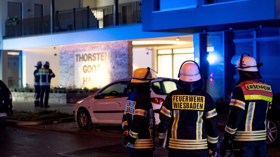 Im Bildvordergrund stehen drei Einsatzkräfte der Feuerwehr in dunkel-gelber Bekleidung und Helmen und blicken in Ricktung eines großen Wohnhauses. Im Bildhintergund unscharf weitere Einsatzkräfte und das Wohnhaus in der Dunkelheit.