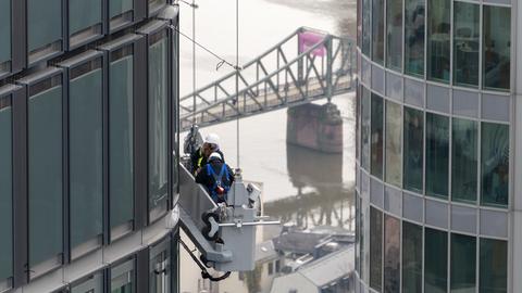 Zwei Fensterputzer arbeiten an der Seite eines Hochhauses in Frankfurt am Main, während im Hintergrund der Eisernen Steg zu sehen ist.