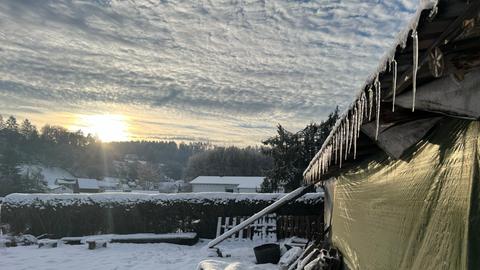 Über einer schneebedeckten Wiese und neben einer Hütte mit Eiszapfen geht die Sonne auf