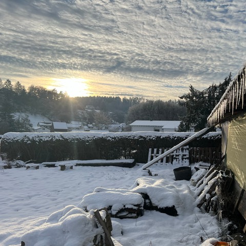 Über einer schneebedeckten Wiese und neben einer Hütte mit Eiszapfen geht die Sonne auf