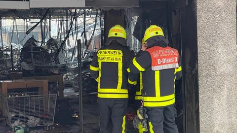 Zwei Feuerwehrleute stehen vor dem ausgebrannten Geschäft neben einem Einkaufswagen, Stahl hängt von der Decke