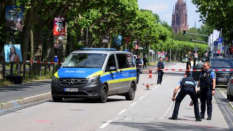 Ein Streifenwagen mit kaputtem Glas steht auf einer Straße, Polizisten machen Markierungen, im Hintergrund der Dom in Frankfurt