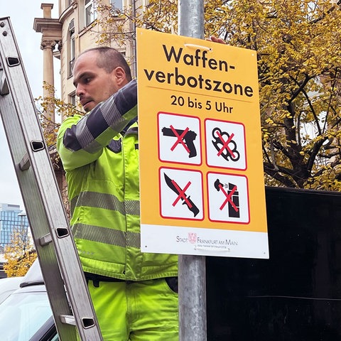 Ein Mann in neonfarbener Arbeitskleidung steht auf einer Leiter und hängt ein Schild mit der Aufschrift 