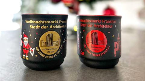 Zwei Tassen für den Weihnachtsmarkt 2024 in Frankfurt. Beide sind schwarz und haben den Aufdruck "Frankfurter Weihnachtsmarkt - Stadt der Architektur - 100 Jahre Neues Frankfurt - Großmarkthalle und EZB" mit einem Bild vom EZB-Hochhaus vor der historischen Großmarkthalle, die eine in Gold, die andere in Rot