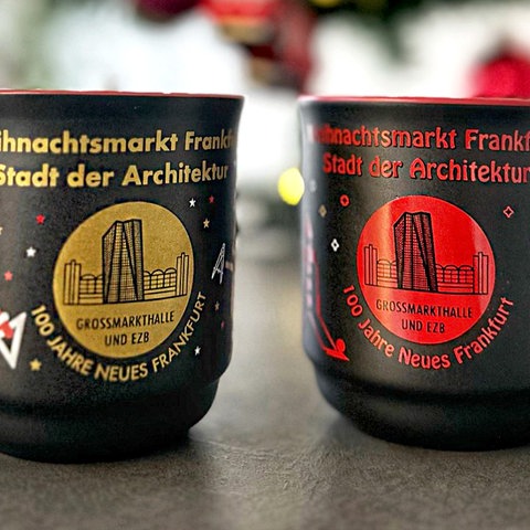 Zwei Tassen für den Weihnachtsmarkt 2024 in Frankfurt. Beide sind schwarz und haben den Aufdruck "Frankfurter Weihnachtsmarkt - Stadt der Architektur - 100 Jahre Neues Frankfurt - Großmarkthalle und EZB" mit einem Bild vom EZB-Hochhaus vor der historischen Großmarkthalle, die eine in Gold, die andere in Rot