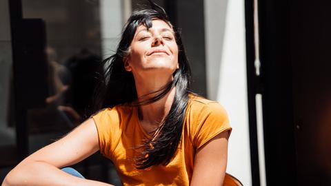 Eine Frau mit langen, dunklen Haaren sitzt im Sonnenlicht und genießt den Moment. Sie trägt ein orangefarbenes T-Shirt. Die Haare wehen leicht im Wind. Der Hintergrund ist unscharf und dunkel, was den Fokus auf die Frau lenkt.