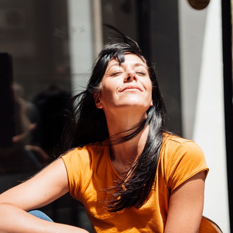 Eine Frau mit langen, dunklen Haaren sitzt im Sonnenlicht und genießt den Moment. Sie trägt ein orangefarbenes T-Shirt. Die Haare wehen leicht im Wind. Der Hintergrund ist unscharf und dunkel, was den Fokus auf die Frau lenkt.