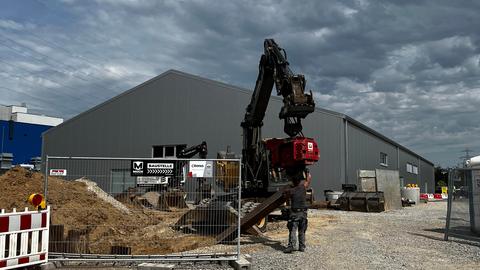 Ein Bauzaun, ein Bagger und ein Bauarbeiter, im Hintergrund die graue Leichtbauhalle, in der der Prozess stattfinden soll