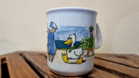 Weiße Tasse mit aufgedrucktem Polizisten, der eine Gans an der Leine führt, daneben aufgemalt ist ein Passant in blauer Jacke und Mütze, der zuschaut