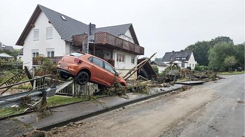 Ein Auto wurde auf einem zerstörten Zaun und Unrat, Leitern, Stöcke, Geäst geschwemmt und scheint in der Luft zu schweben. Im Hintergund ein Wohnhaus. Der Bereich vor dem Haus ist angereichert mit Geäst, Steinplatten ung angeschwemmten Zeugs. 