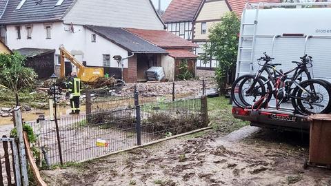 Foto eines Wohnmobils von hinten, das in einer Einfahrt steht, die überflutet von Schlamm ist. Auch die umgebenden Gärten sind voller Schlamm, Wasser und Zerstörung und ein Feuerwehrmann steht darin.