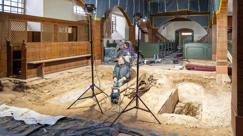 Kircheninnenraum mit Ausgrabungen im Kirchenboden: zwei tiefe Gräber. Davor zwei Leuchten und zwei Menschen darin sitzend.