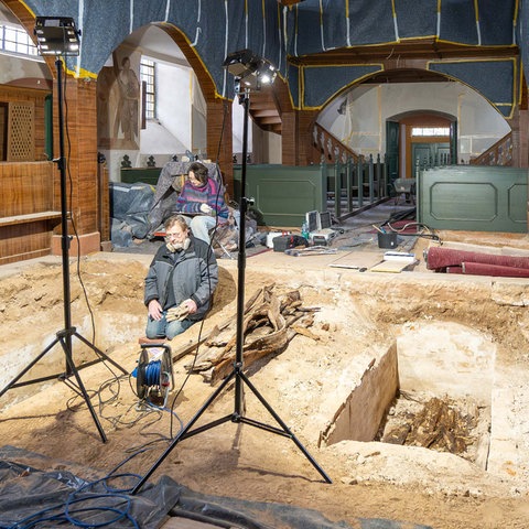 Kircheninnenraum mit Ausgrabungen im Kirchenboden: zwei tiefe Gräber. Davor zwei Leuchten und zwei Menschen darin sitzend.