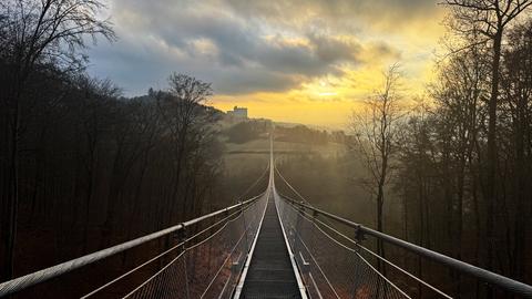 Fotografischer Blick auf eine Fußgängerhängebrücke und ins Tal daneben bei Sonnenaufgang.