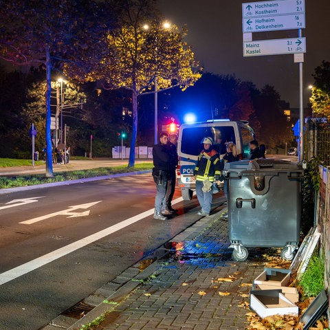 Feuerwehrleute und Polizisten an einer Straße mit Mülltonne im Vordergrund