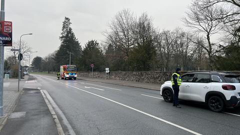 Ein Polizist steht neben einem weißen Auto auf einer Straße und spricht mit dem Fahrer. Auf der Straße steht auch ein Feuerwehrfahrzeug mit blinkenden Lichtern und einem Feuerwehrmann daneben. Ansonsten ist die Straße leer. Im Hintergrund sind Bäume und Häuser zu sehen. 