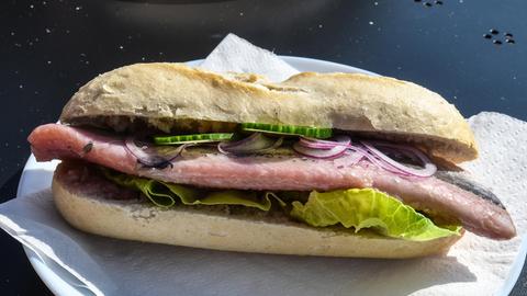 Ein Sandwich mit Fischfilet, grüner Salat, Gurkenscheiben und roten Zwiebelringen auf einem Baguette-Brötchen. Es liegt auf einem weißen Teller mit einer Serviette darunter.