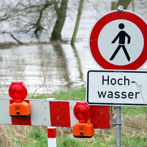 Ein über die Ufer getretener Fluss. Davor ein Schild "Hochwasser" und ein rot-weißes Absperrgestell.