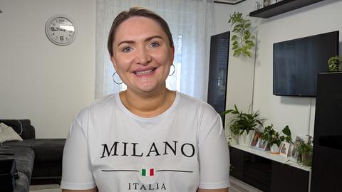 Valentina Simin sitzt in ihrem Wohnzimmer. Sie hat ihre blonden Haare zu einem Zopf gebunden, auf ihrem weißen T-Shirt steht "Milano - italia".