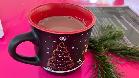 Eine schwarz-rote Tasse mit Kakao steht auf einem Tisch des Frankfurter Weihnachtsmarkts. Neben der Tasse liegt ein Tannenzweig.